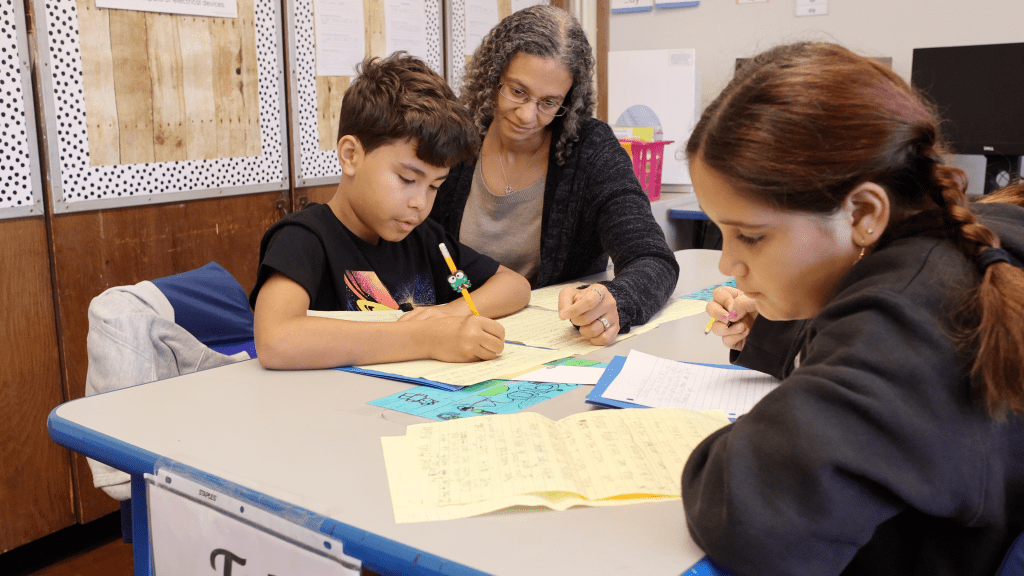 teacher and student working together at a table with another student working independently